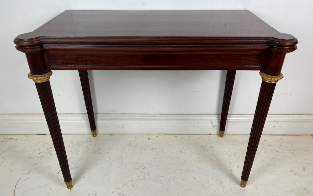 A polished dark mahogany fold-over card table featuring tapered fluted legs topped with gilt bronze acanthus collars.