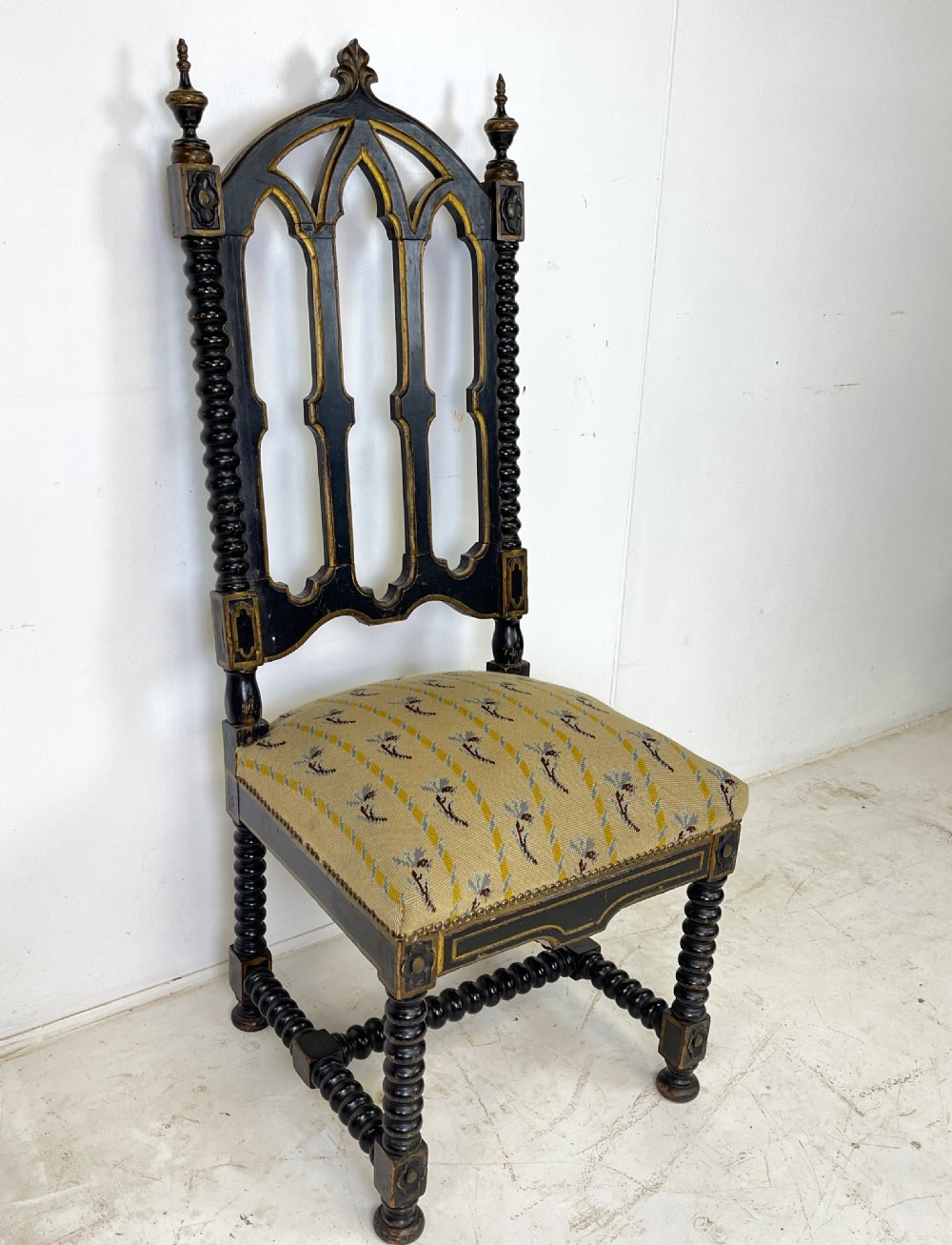A Victorian Gothic Revival side chair featuring an ebonised frame with lancet arch tracery, bobbin turned legs, and patterned cream upholstery with brass stud details. View V
