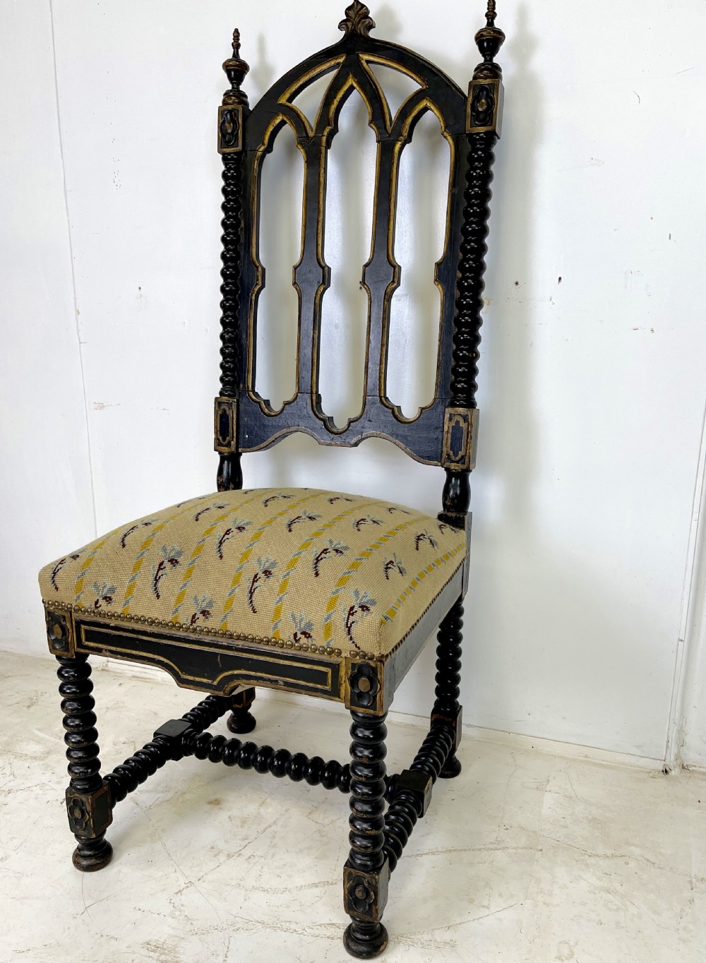 A Victorian Gothic Revival side chair featuring an ebonised frame with lancet arch tracery, bobbin turned legs, and patterned cream upholstery with brass stud details. View VI