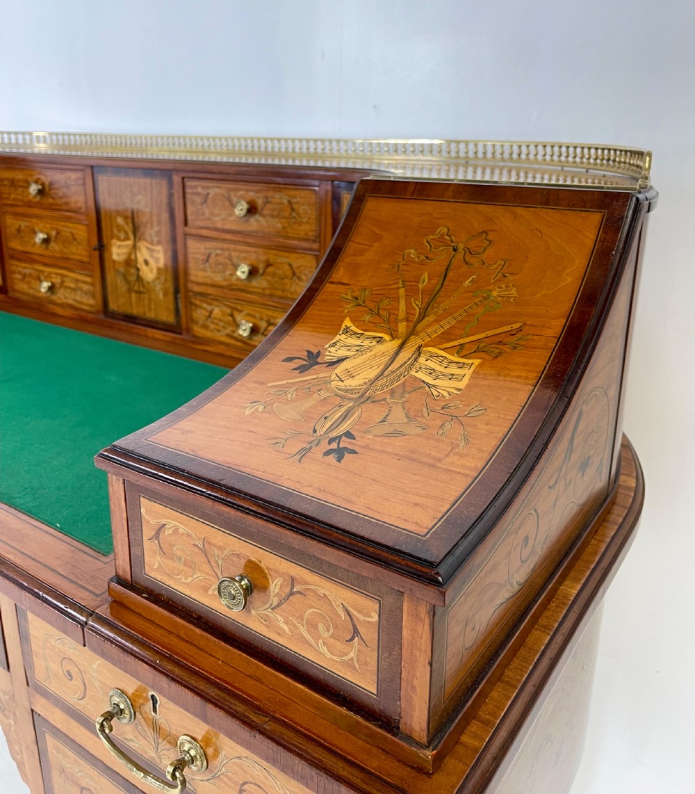 An Edwardian satinwood Carlton House desk with floral marquetry inlay, a green leather writing surface, multiple drawers, and a brass gallery top. View V
