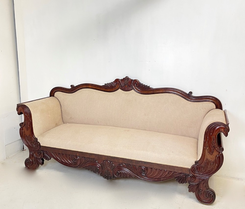 A large antique mahogany sofa featuring intricately hand-carved acanthus scrollwork along the apron and arms, upholstered in a light cream textured fabric.
