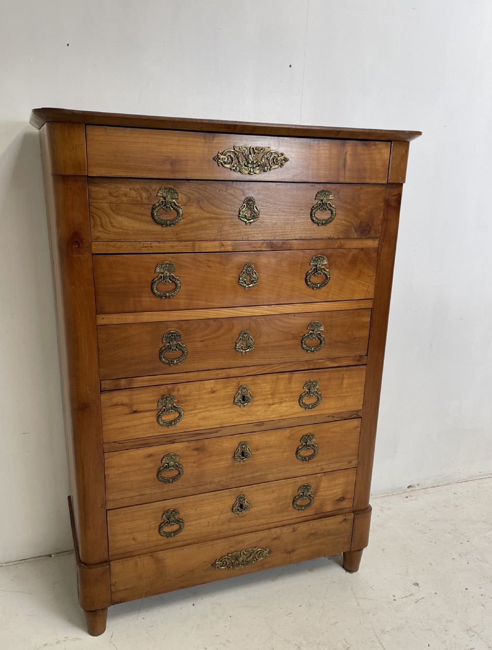 A tall French cherrywood semainier featuring seven drawers with brass drop handles and decorative keyhole escutcheons. View IX