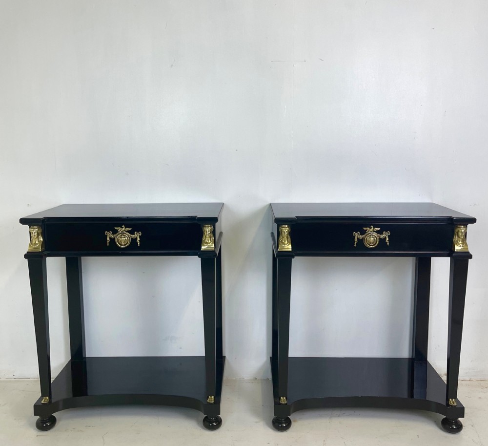 A pair of ebonised black side tables featuring single drawers, lower shelves, and gilt brass mounts on square legs.