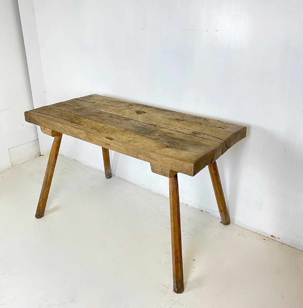 An antique rustic wooden work table featuring a thick timber top supported by four splayed cylindrical legs with visible wear and grain. View III
