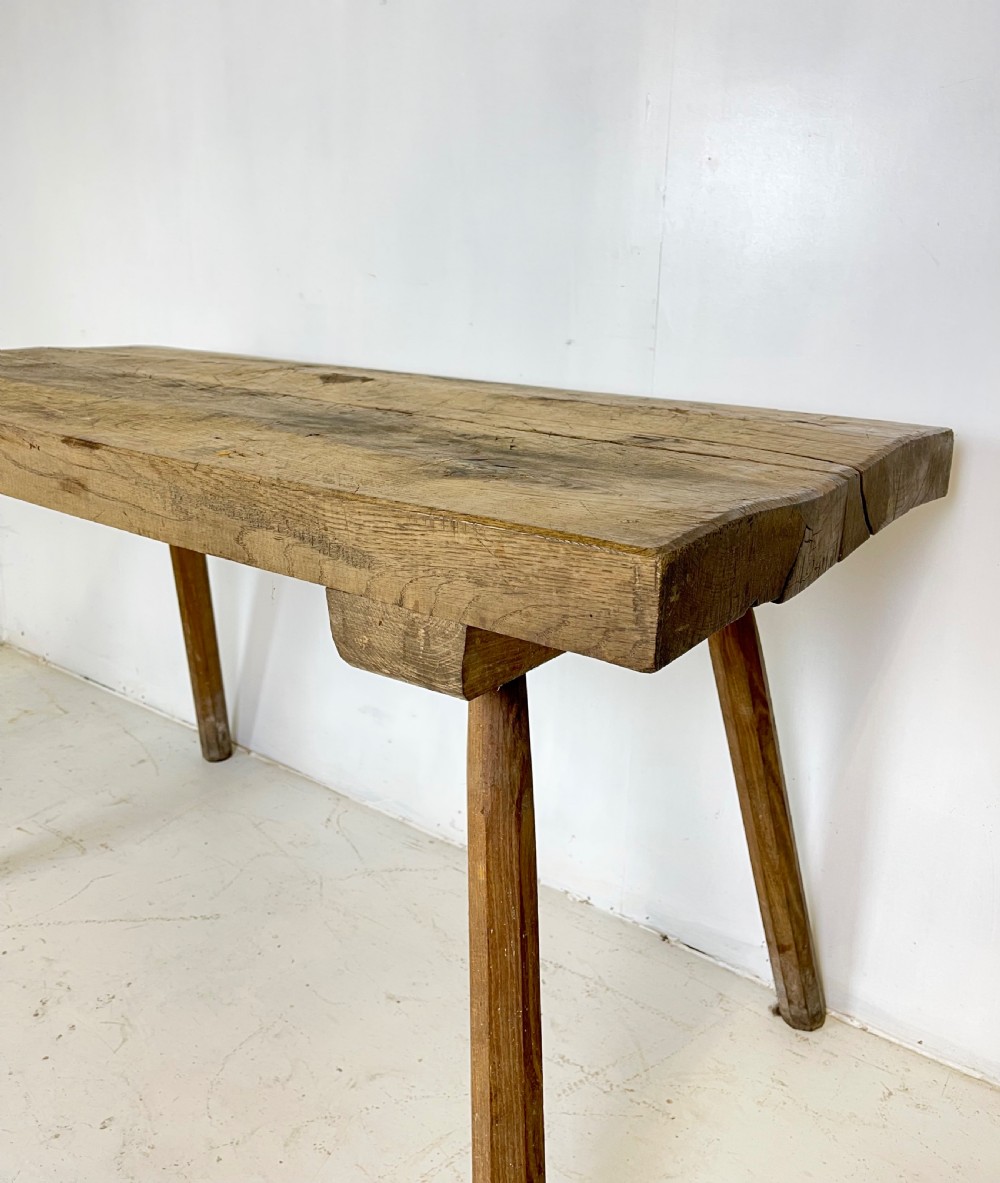 An antique rustic wooden work table featuring a thick timber top supported by four splayed cylindrical legs with visible wear and grain. View IV