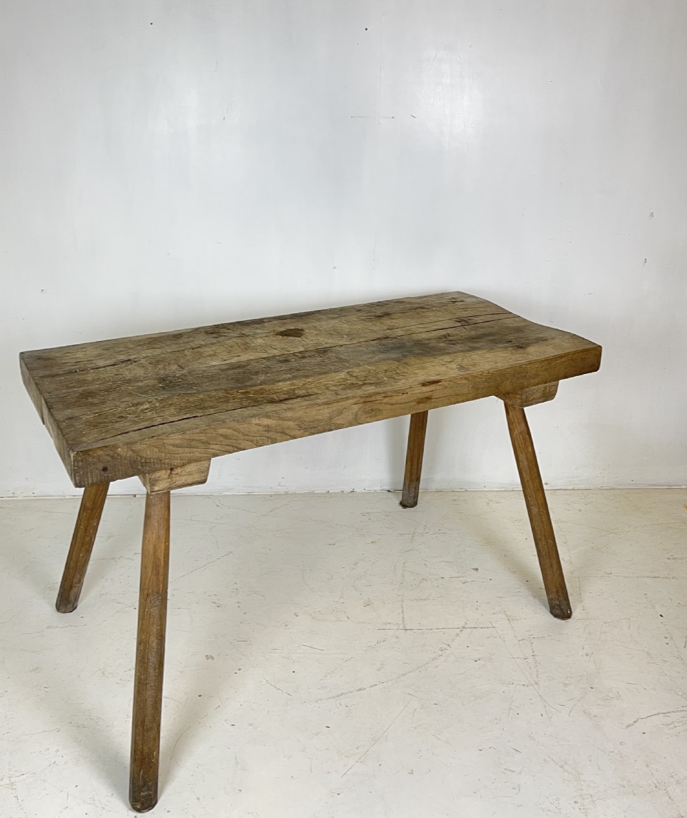 An antique rustic wooden work table featuring a thick timber top supported by four splayed cylindrical legs with visible wear and grain. View VI
