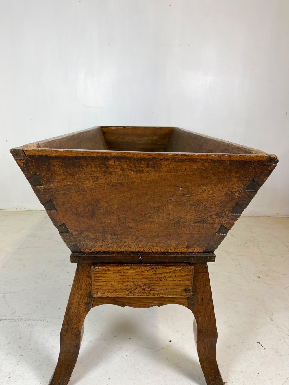 An antique French walnut dough bin featuring a flat overhanging top, sloped timber sides, and a scalloped apron resting on short legs. View VII