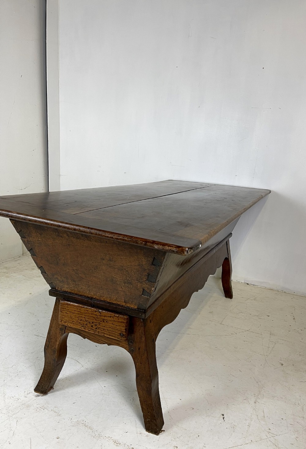 An antique French walnut dough bin featuring a flat overhanging top, sloped timber sides, and a scalloped apron resting on short legs. View XI