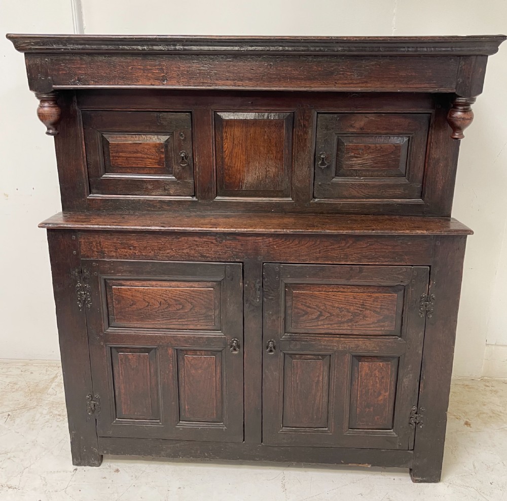 A dark oak joined court cupboard featuring a recessed upper section with geometric panelled doors and turned drop finials above a two-door base.