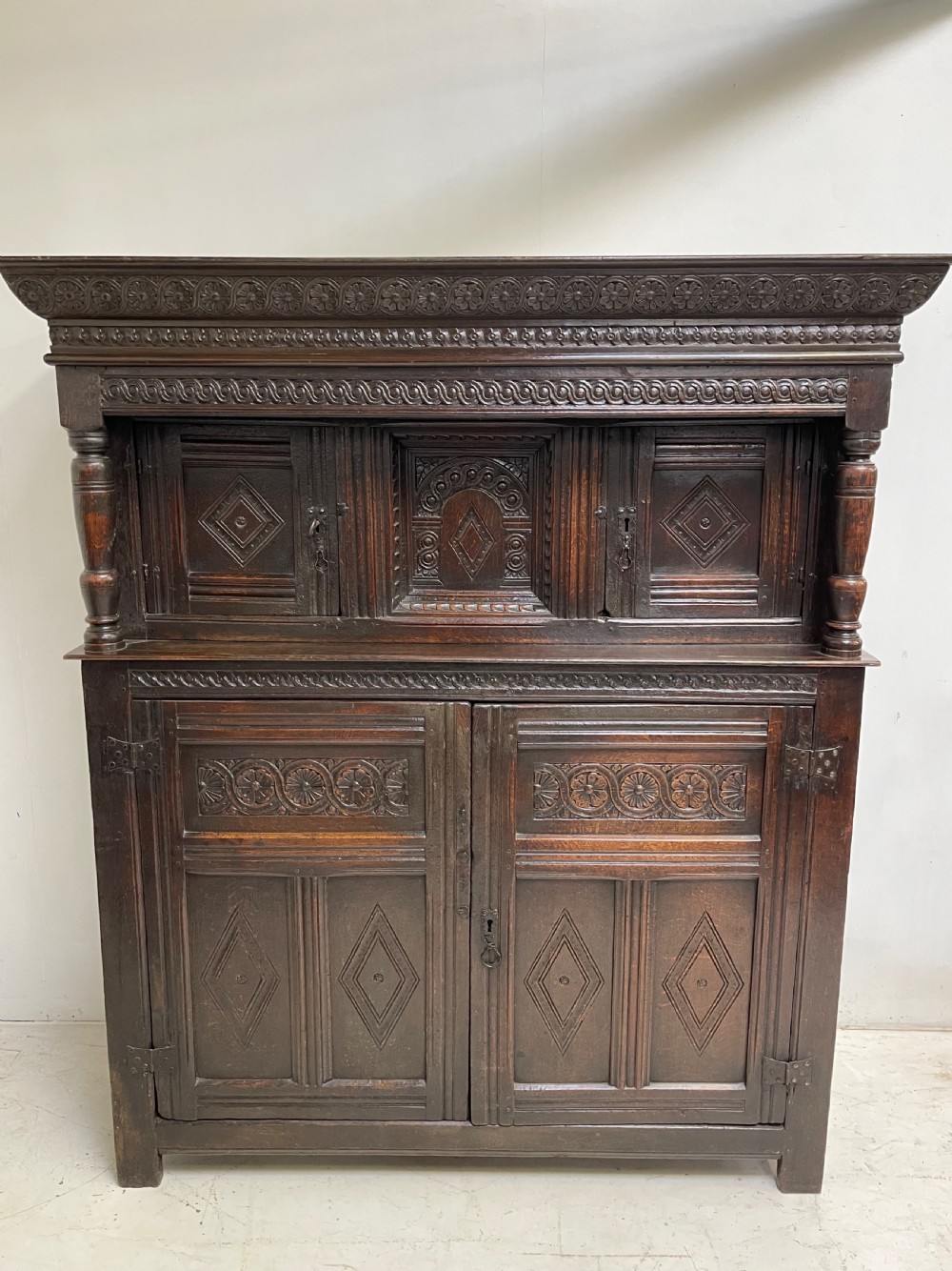 A dark carved oak court cupboard featuring upper and lower double doors with geometric diamond panels, guilloche frieze carving, and turned side supports.