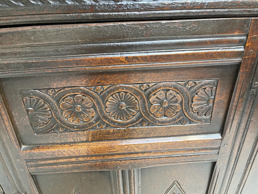 A dark carved oak court cupboard featuring upper and lower double doors with geometric diamond panels, guilloche frieze carving, and turned side supports. View II