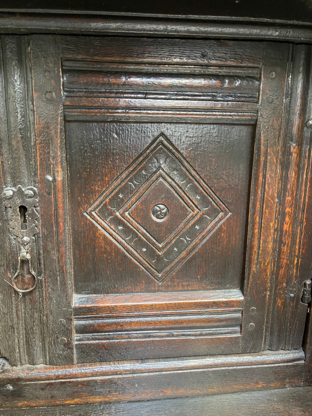 A dark carved oak court cupboard featuring upper and lower double doors with geometric diamond panels, guilloche frieze carving, and turned side supports. View III