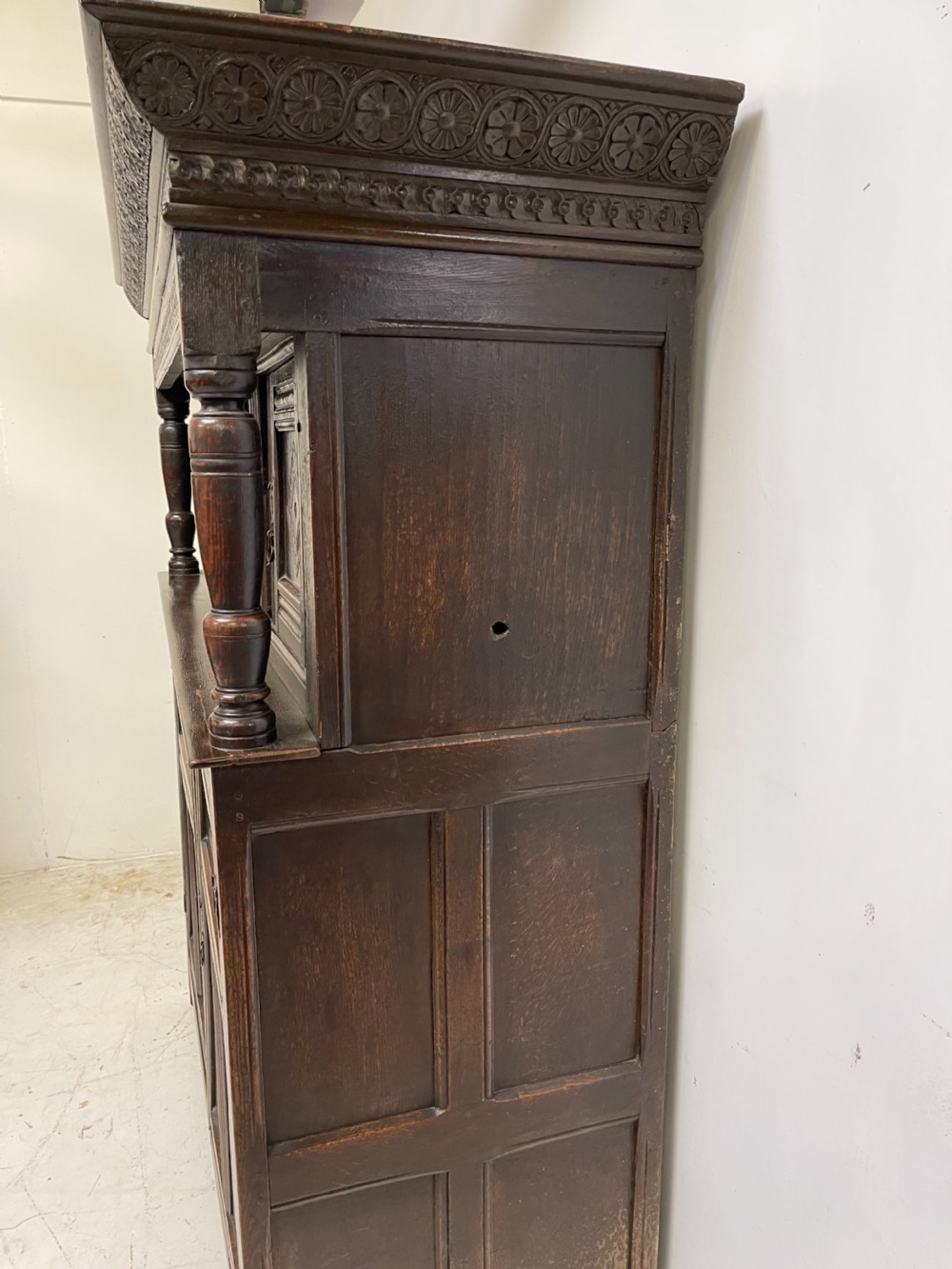 A dark carved oak court cupboard featuring upper and lower double doors with geometric diamond panels, guilloche frieze carving, and turned side supports. View V