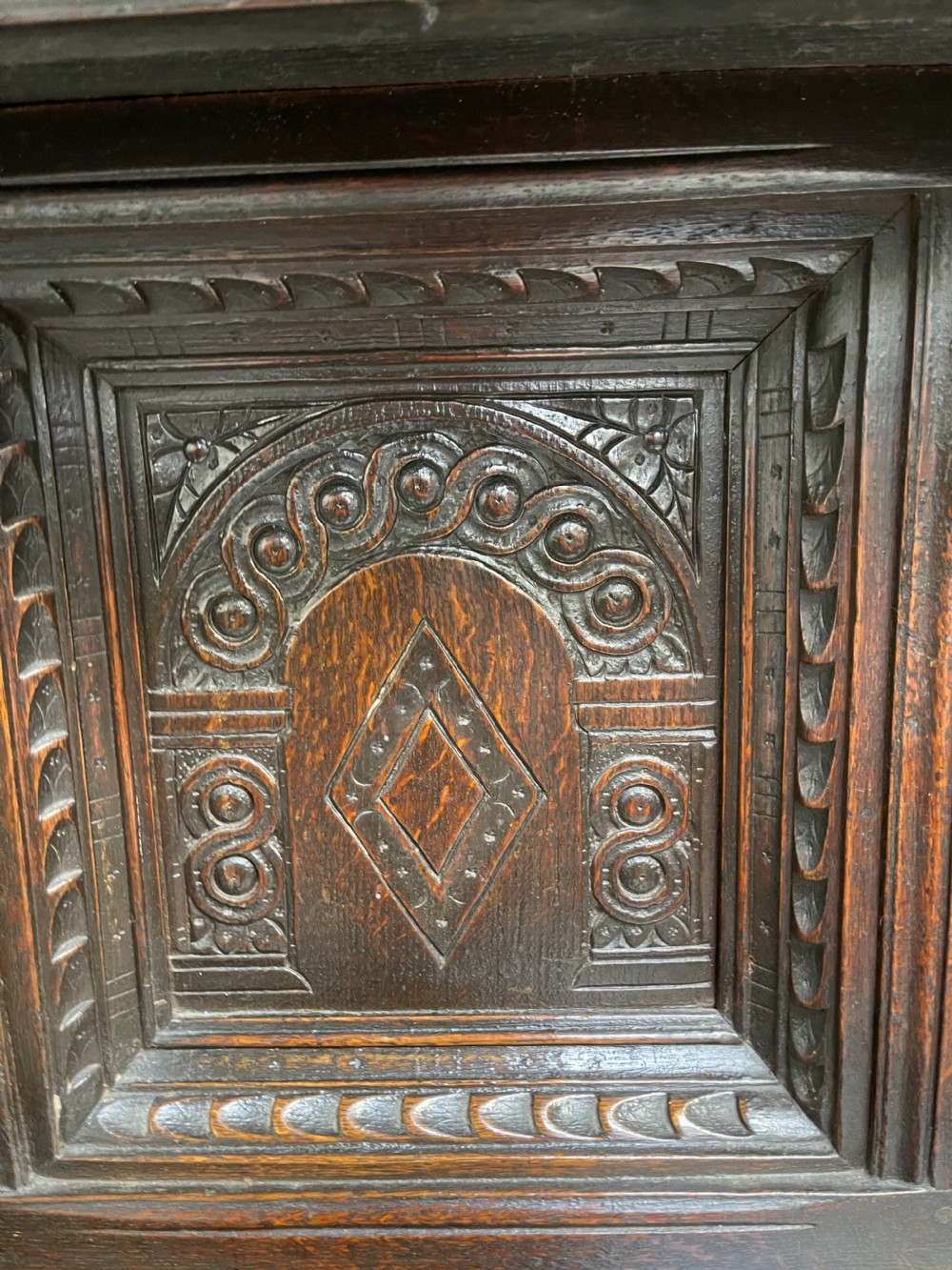 A dark carved oak court cupboard featuring upper and lower double doors with geometric diamond panels, guilloche frieze carving, and turned side supports. View VI