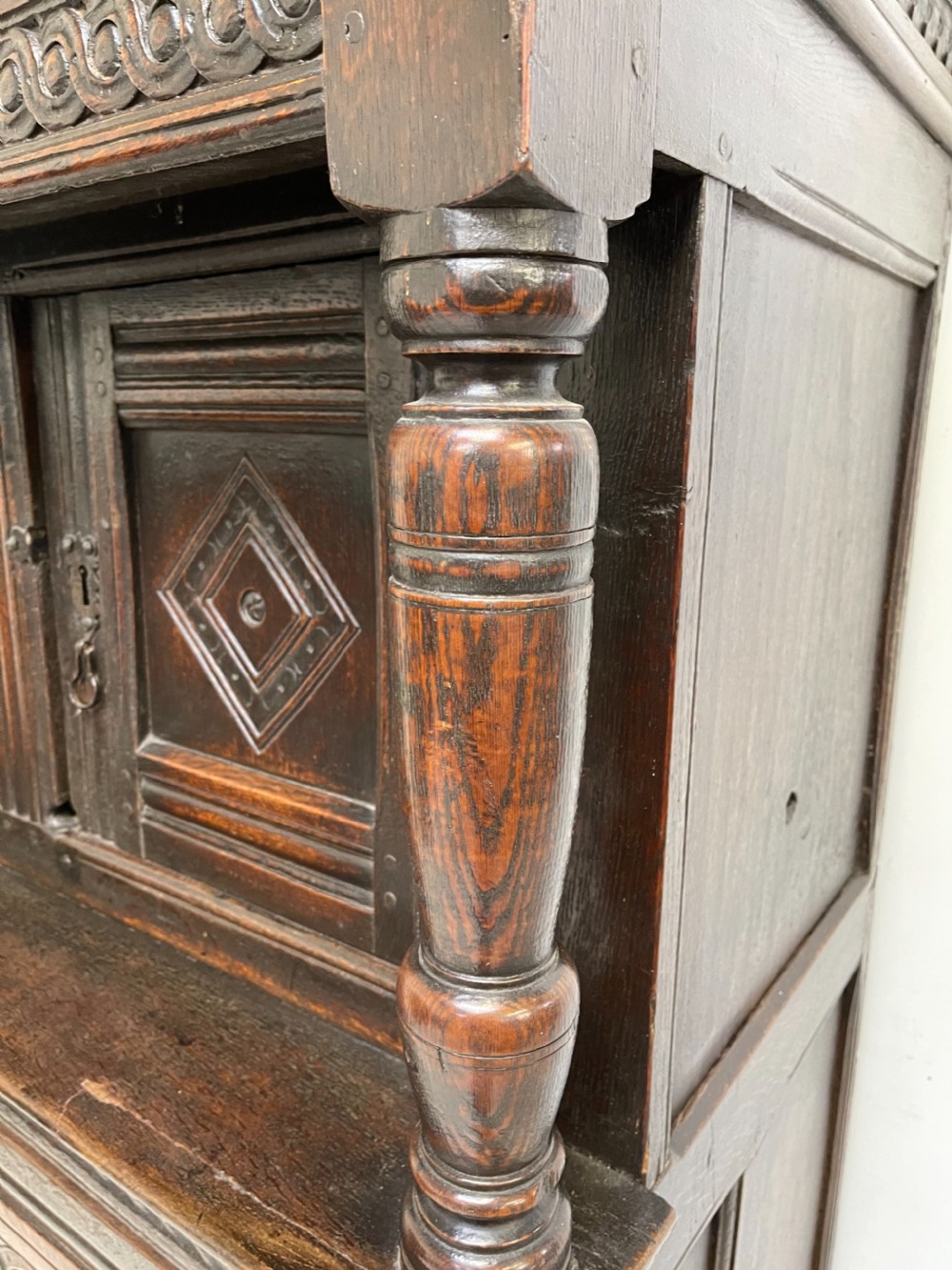 A dark carved oak court cupboard featuring upper and lower double doors with geometric diamond panels, guilloche frieze carving, and turned side supports. View IX