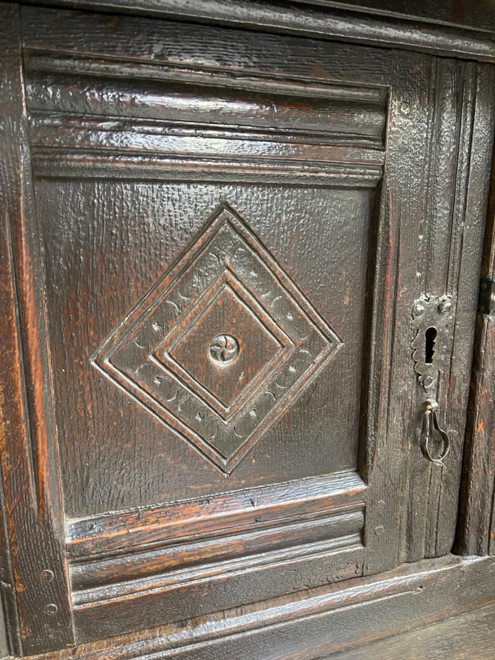 A dark carved oak court cupboard featuring upper and lower double doors with geometric diamond panels, guilloche frieze carving, and turned side supports. View XIV