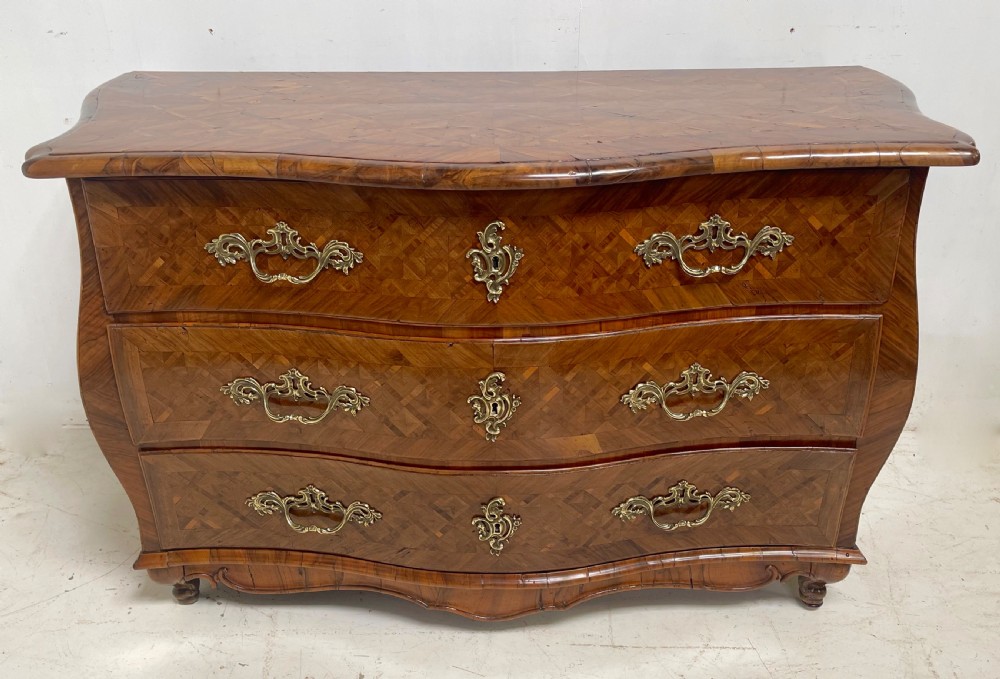 An 18th-century French bombe commode featuring three drawers with geometric parquetry inlay, a serpentine silhouette, and ornate brass handles.