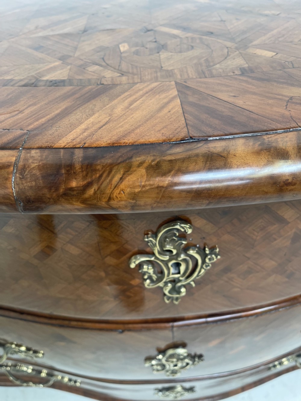 An 18th-century French bombe commode featuring three drawers with geometric parquetry inlay, a serpentine silhouette, and ornate brass handles. View V