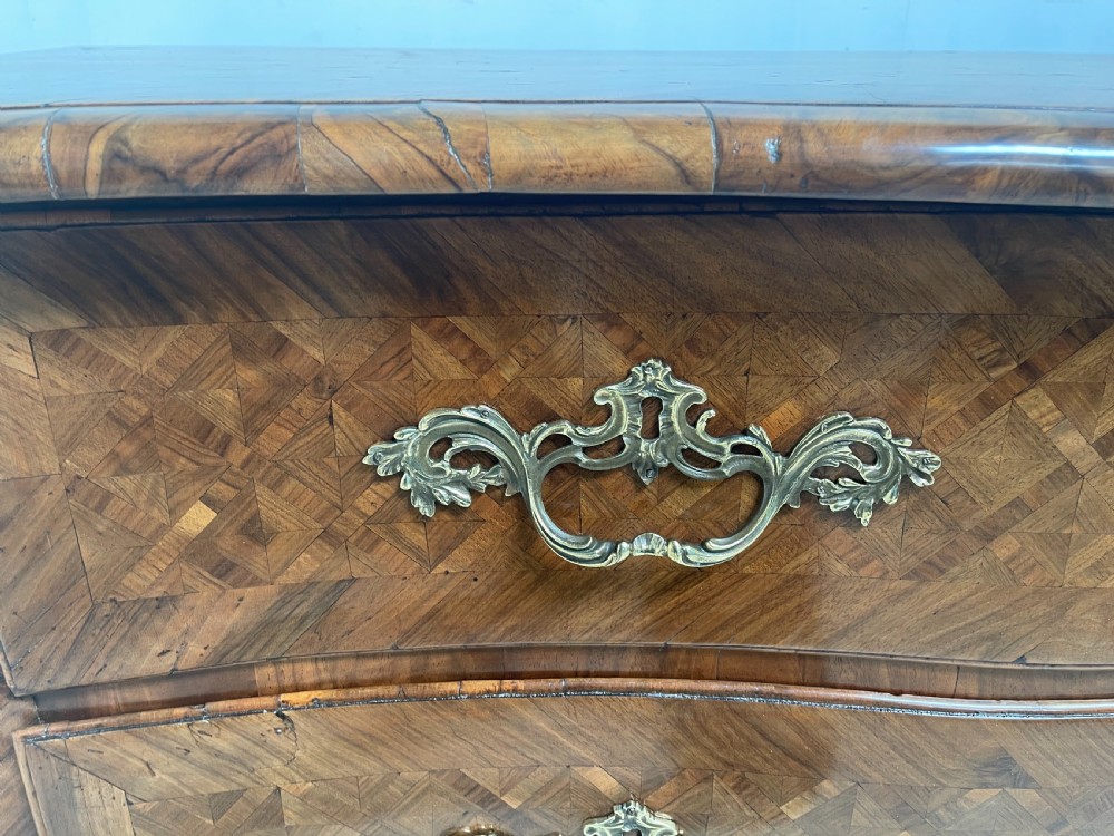 An 18th-century French bombe commode featuring three drawers with geometric parquetry inlay, a serpentine silhouette, and ornate brass handles. View VI