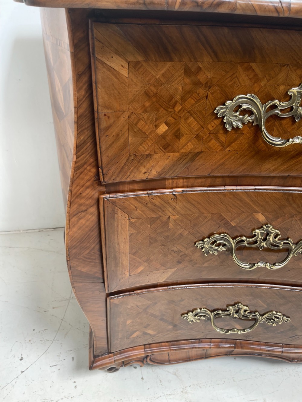 An 18th-century French bombe commode featuring three drawers with geometric parquetry inlay, a serpentine silhouette, and ornate brass handles. View IX