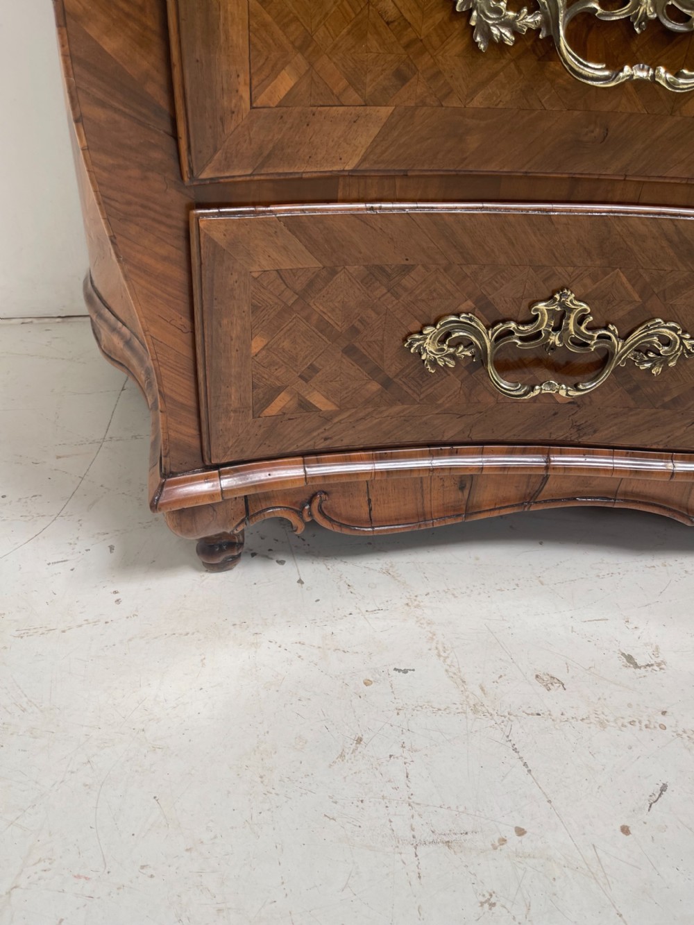 An 18th-century French bombe commode featuring three drawers with geometric parquetry inlay, a serpentine silhouette, and ornate brass handles. View X