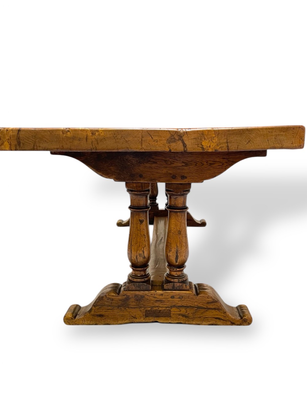 A solid warm brown oak refectory dining table featuring a thick rectangular top supported by double turned columns and a central stretcher. View VII