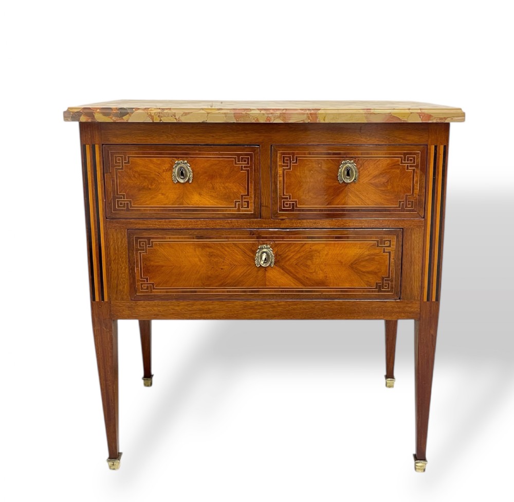 A Louis XVI style wooden commode featuring geometric parquetry inlay, a variegated marble top, three drawers with brass escutcheons, and tapered square legs.
