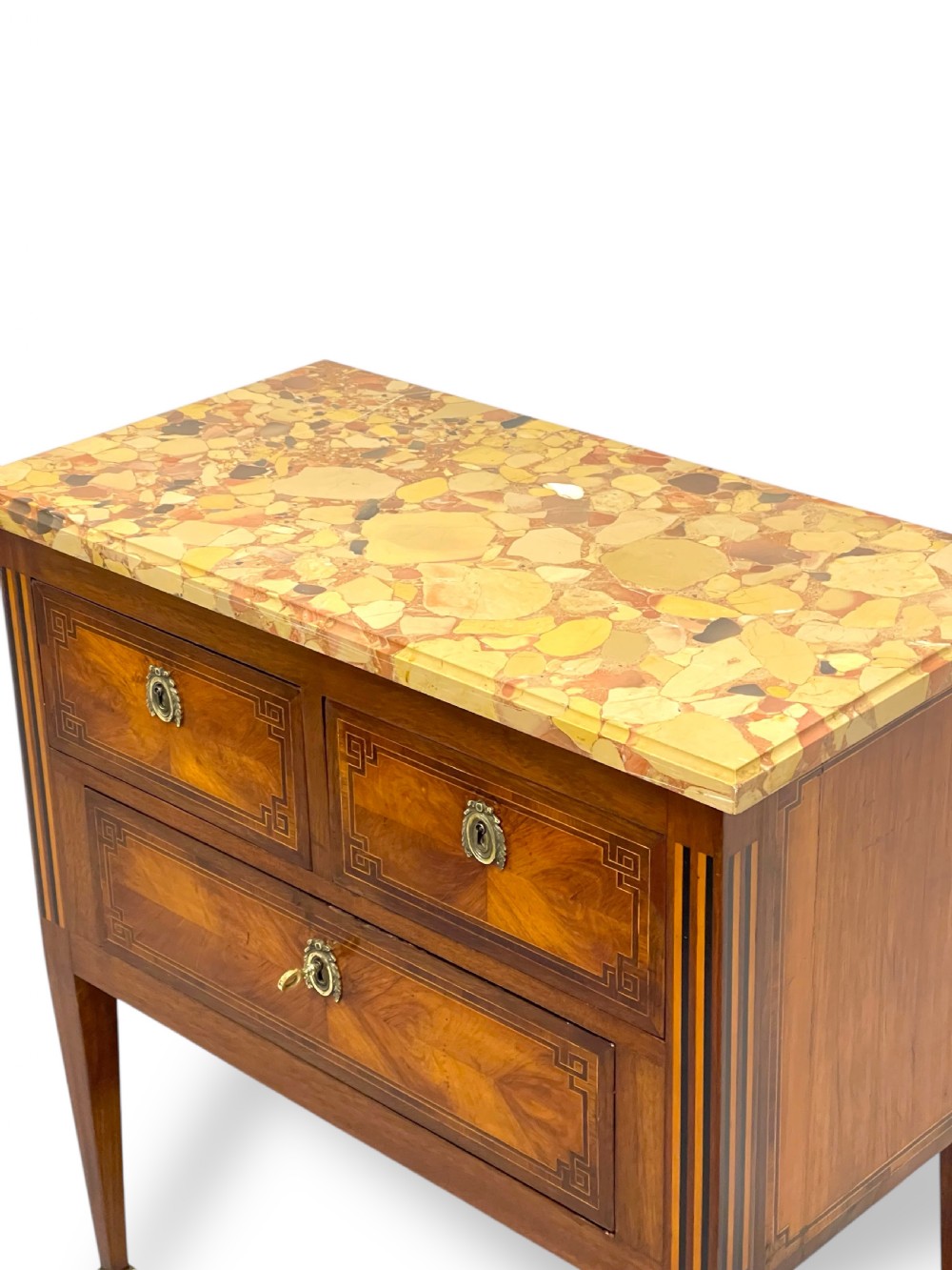 A Louis XVI style wooden commode featuring geometric parquetry inlay, a variegated marble top, three drawers with brass escutcheons, and tapered square legs. View V