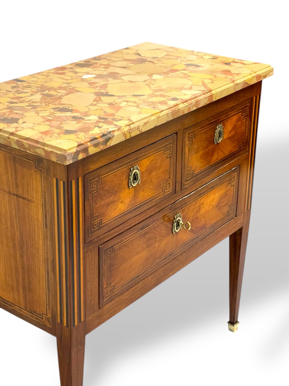 A Louis XVI style wooden commode featuring geometric parquetry inlay, a variegated marble top, three drawers with brass escutcheons, and tapered square legs. View VIII