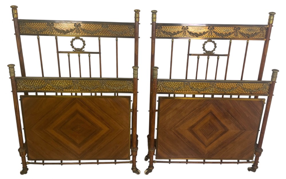 A pair of Edwardian brass and walnut single bed frames featuring diamond veneer panels and brass swag detailing.