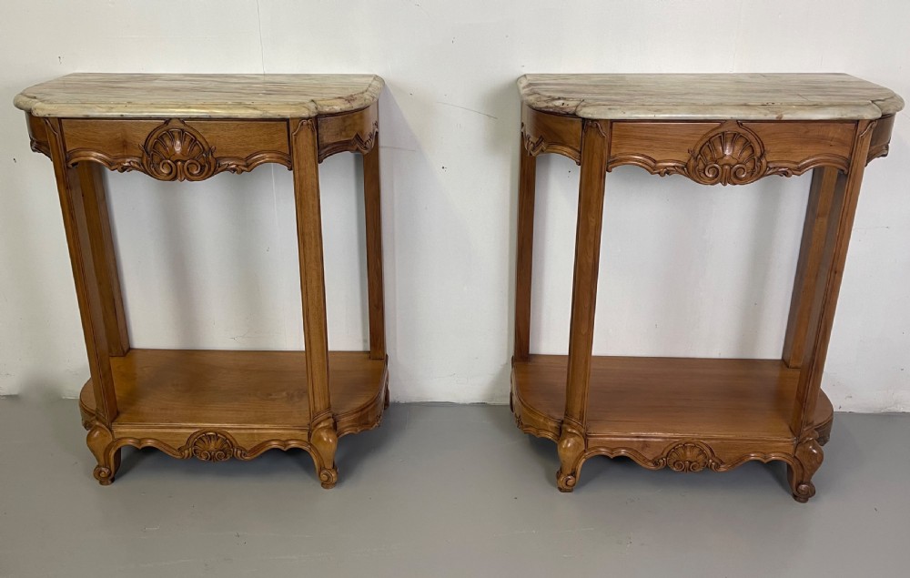 A pair of French Louis XV style walnut console tables featuring shaped marble tops, carved shell aprons, lower shelves, and cabriole legs.