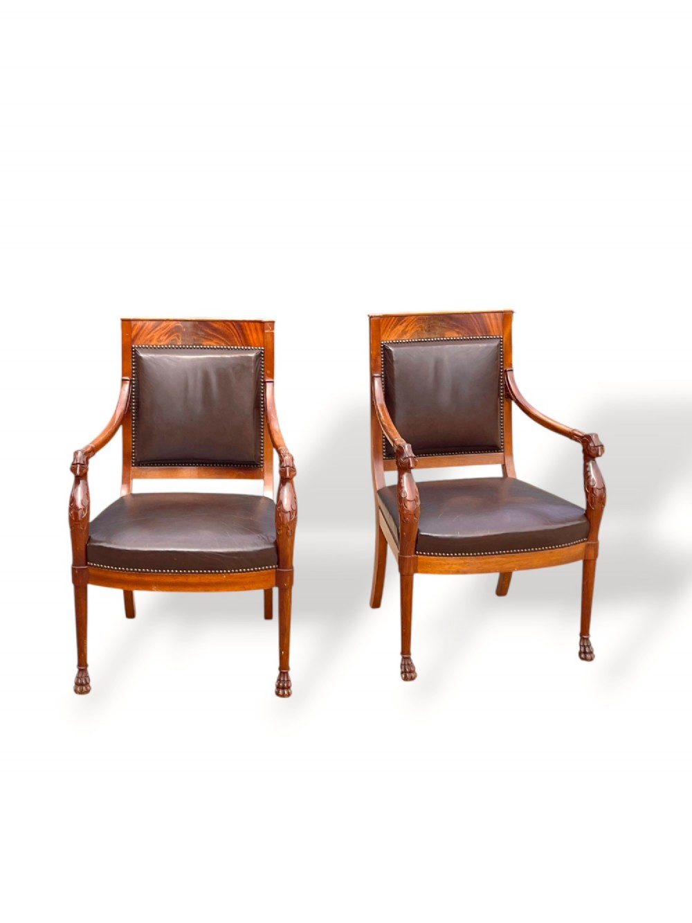 A pair of twentieth-century French Empire style armchairs featuring carved mahogany frames, dark brown leather upholstery, and brass stud trim.