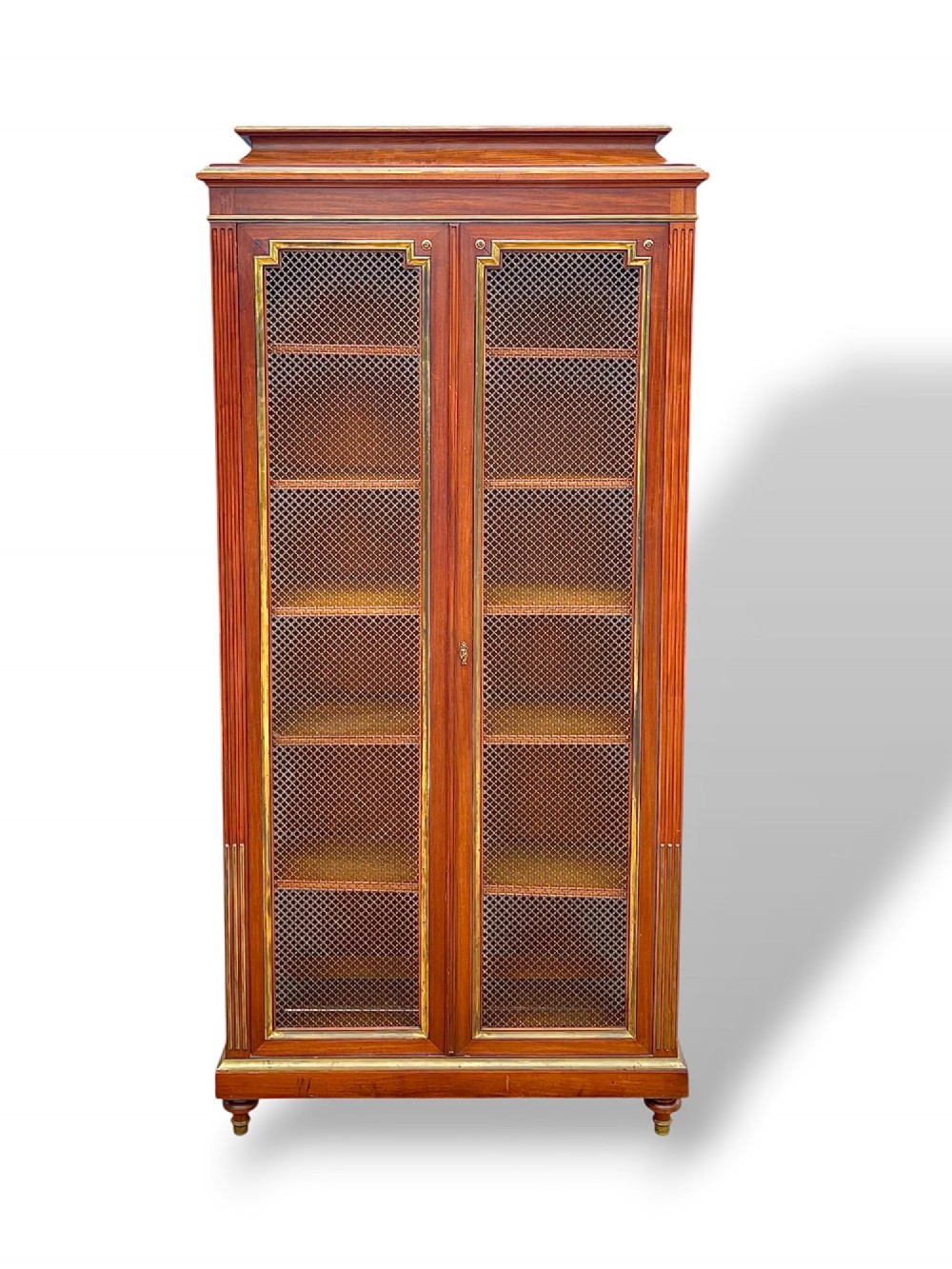 A tall French Empire style mahogany two-door bookcase featuring brass wire mesh door panels and fluted side columns.