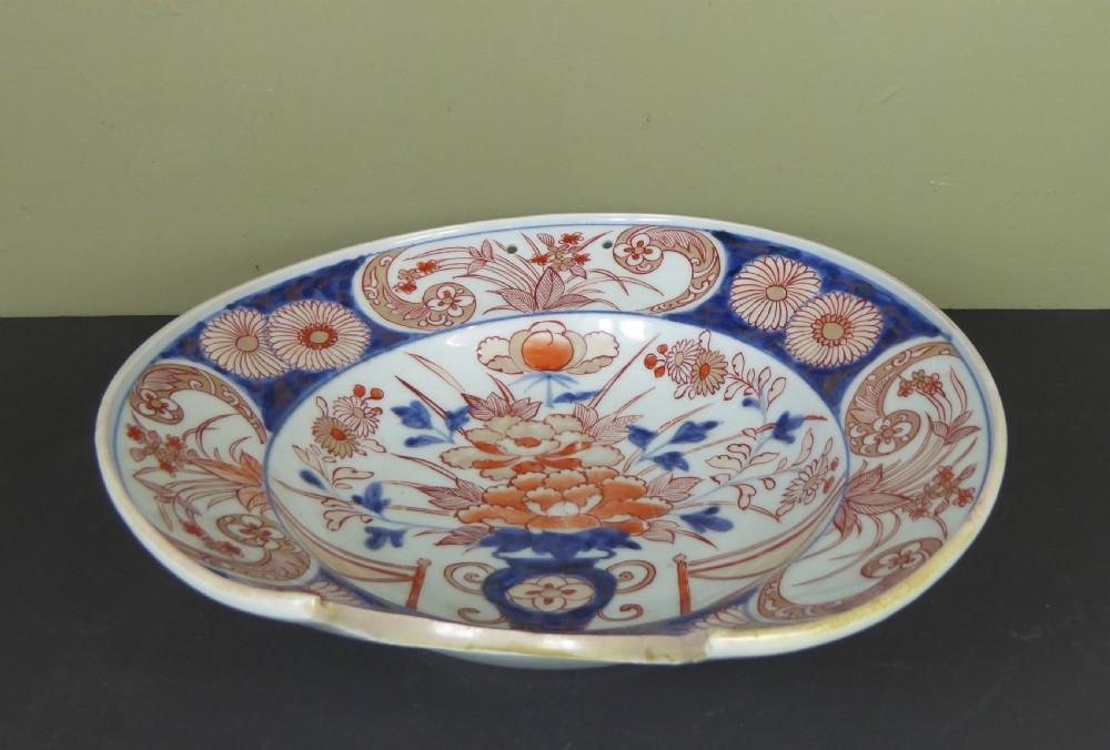 An 18th-century Imari porcelain shaving bowl decorated with hand-painted blue and iron-red floral motifs, featuring a crescent cut-out along the rim.