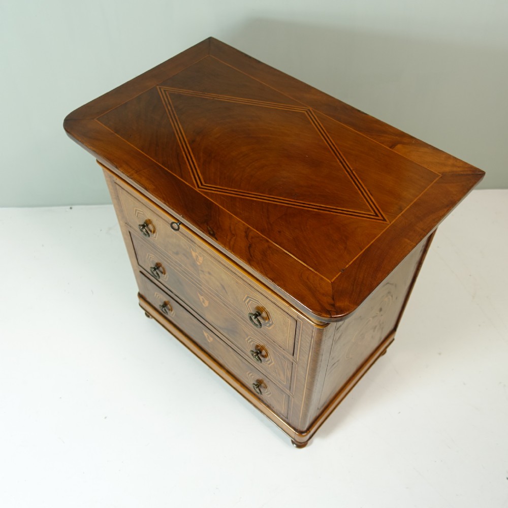 A 19th-century miniature wooden chest of drawers featuring three inlaid drawers, octagonal ring handles, shield escutcheons, and small turned feet. View VI