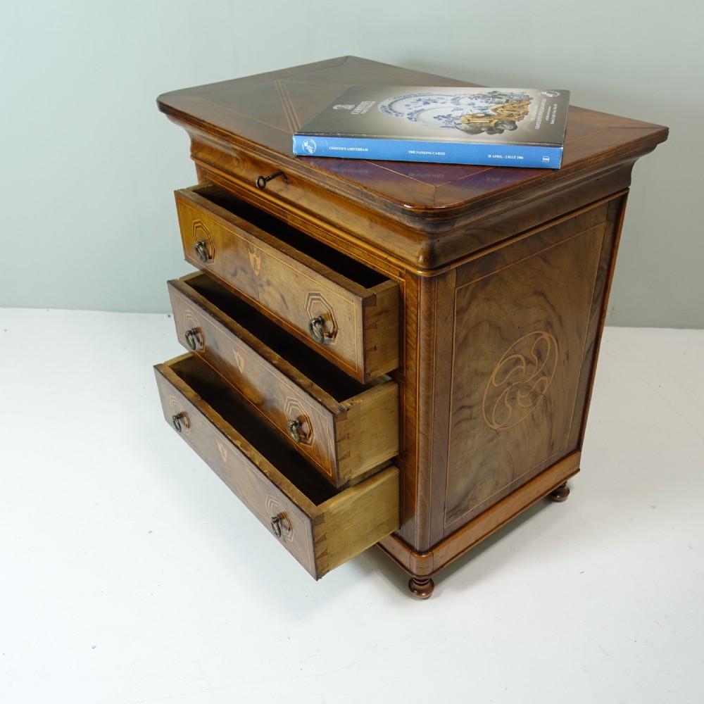 A 19th-century miniature wooden chest of drawers featuring three inlaid drawers, octagonal ring handles, shield escutcheons, and small turned feet. View VII