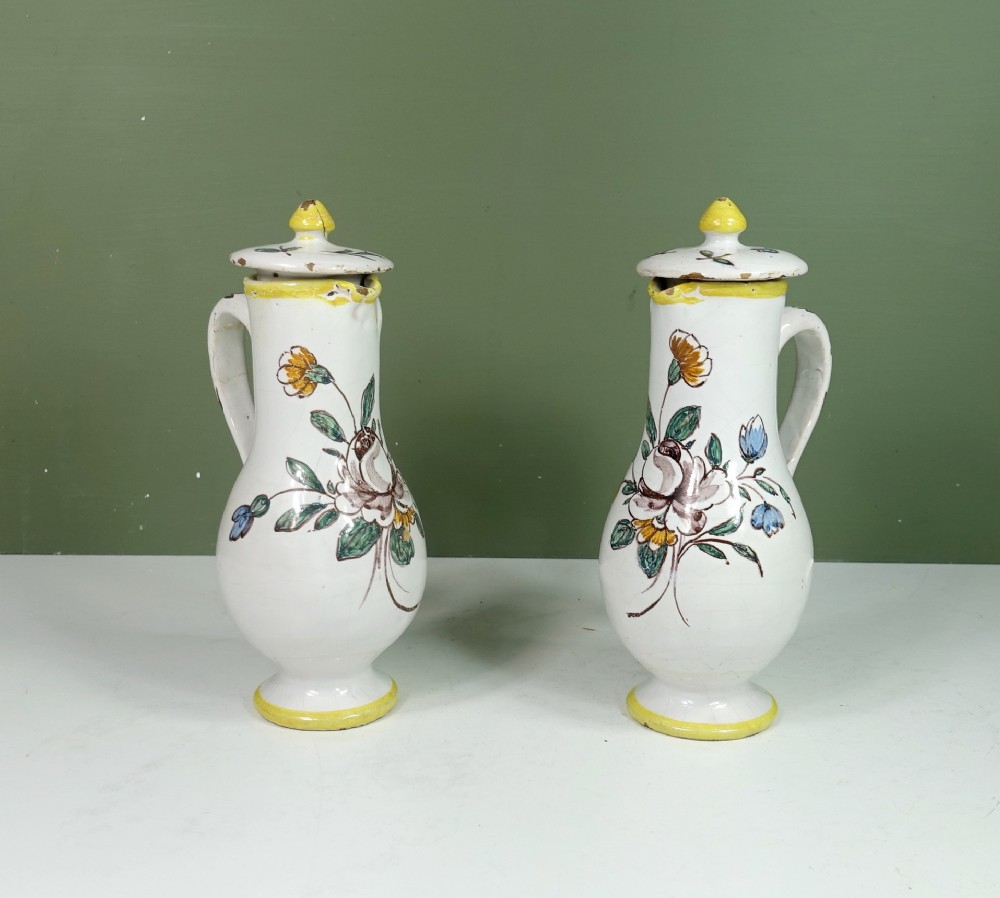 A pair of 19th-century white tin-glazed earthenware jugs featuring hand-painted floral motifs in yellow, green, brown, and blue with matching lidded tops.