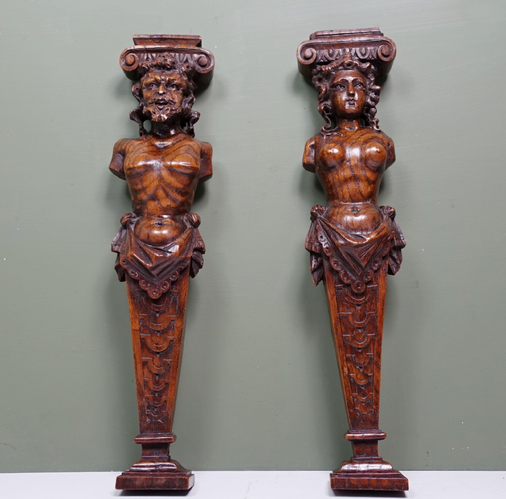 A pair of early nineteenth-century carved chestnut herms depicting male and female busts with Ionic scroll capitals above tapered, scale-carved bases.
