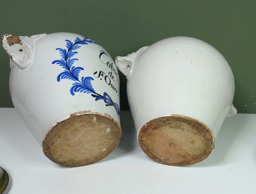 A pair of 18th-century white tin-glazed earthenware tobacco jars featuring blue hand-painted wreaths, inscriptions, molded handles, and stepped brass lids. View V