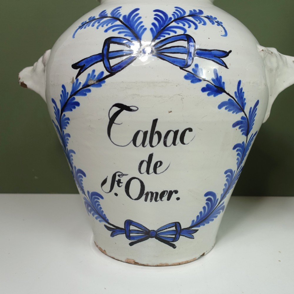 A pair of 18th-century white tin-glazed earthenware tobacco jars featuring blue hand-painted wreaths, inscriptions, molded handles, and stepped brass lids. View VI
