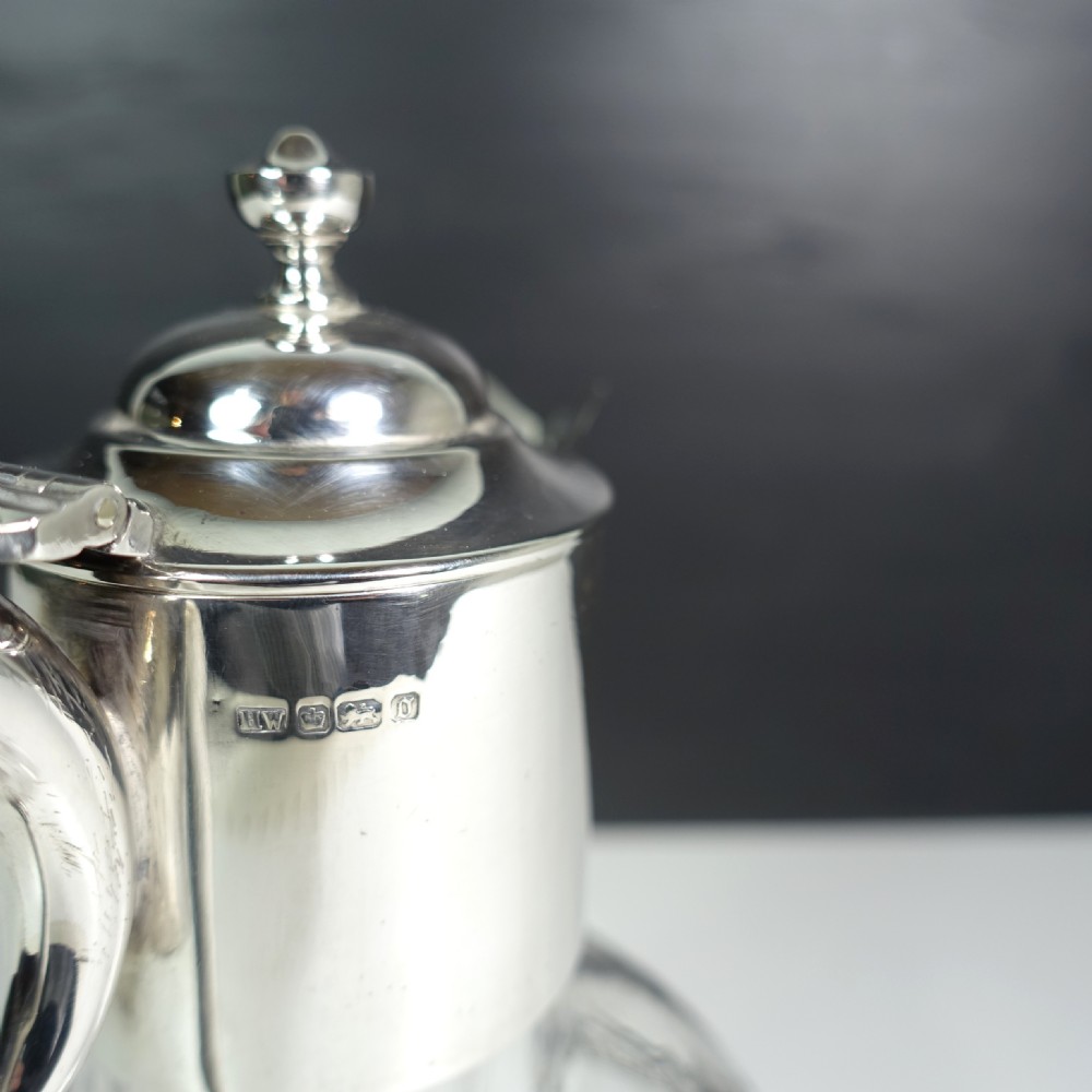 A silver-mounted cut glass claret jug featuring a polished metal spout, handle, hinged lid, and intricate geometric diamond-cut patterns around the clear glass body. View VI