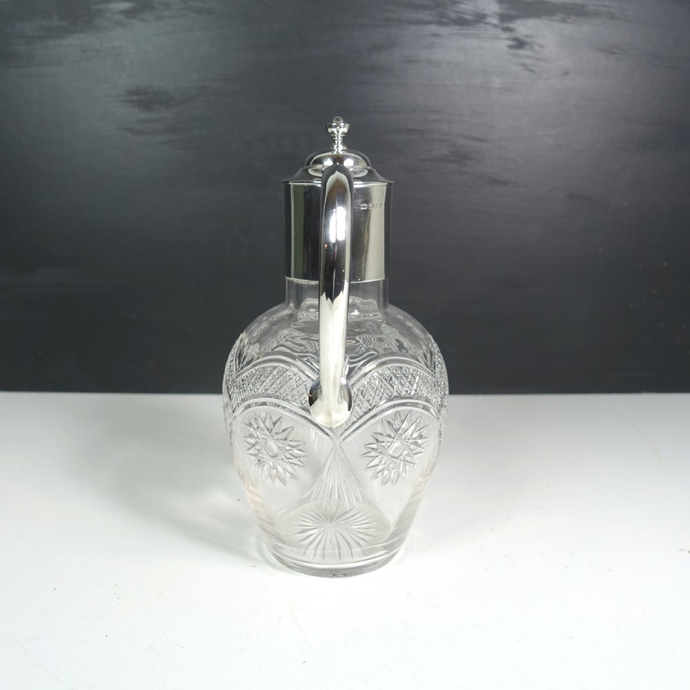 A silver-mounted cut glass claret jug featuring a polished metal spout, handle, hinged lid, and intricate geometric diamond-cut patterns around the clear glass body. View VII