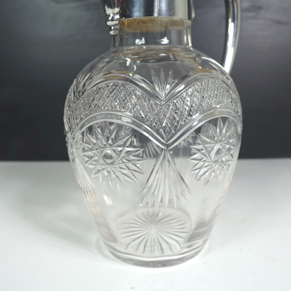 A silver-mounted cut glass claret jug featuring a polished metal spout, handle, hinged lid, and intricate geometric diamond-cut patterns around the clear glass body. View VIII