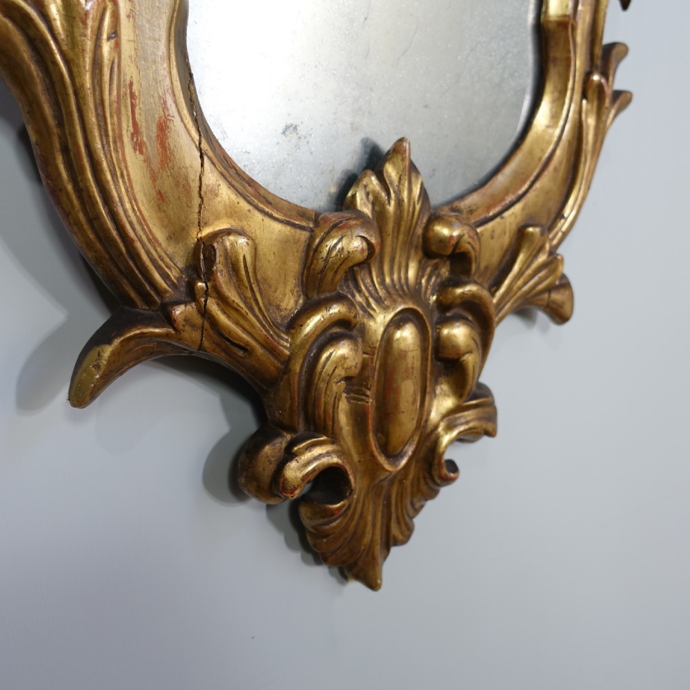 An 18th-century carved giltwood wall mirror featuring elaborate scrolling acanthus leaves, a central crest, and foxed mirror glass. View IV