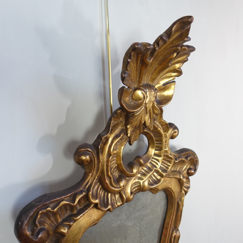 An 18th-century carved giltwood wall mirror featuring elaborate scrolling acanthus leaves, a central crest, and foxed mirror glass. View V
