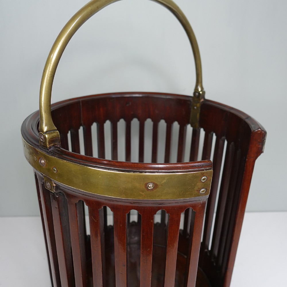 A circular Georgian mahogany slatted plate bucket with brass bound rim and base, a brass swing handle, and a cut-out front opening containing dinner plates. View III