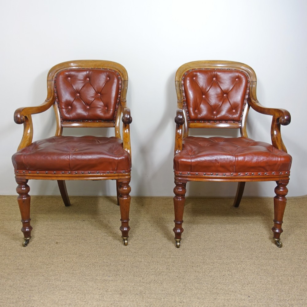 A pair of 19th-century mahogany armchairs upholstered in deep-buttoned reddish-brown leather with brass stud detail and turned front legs on castors.