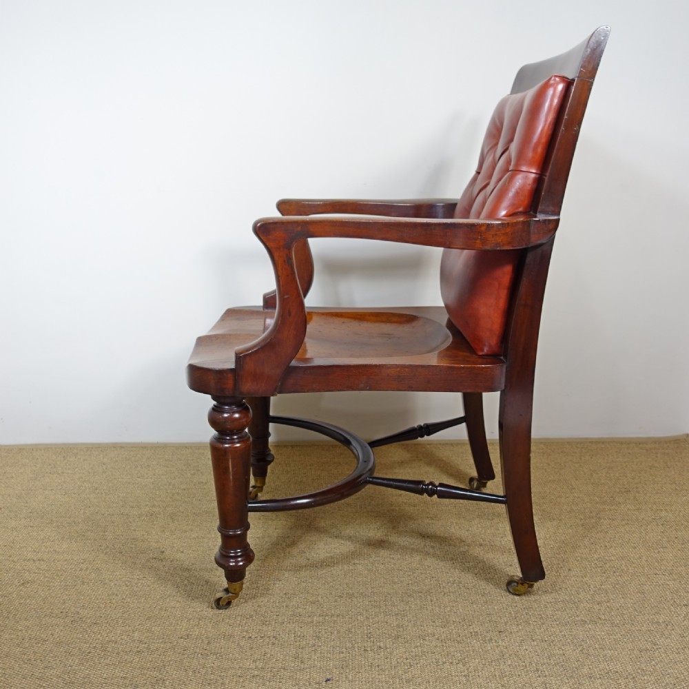 A 19th-century mahogany open armchair featuring a red buttoned-leather backrest, wooden seat, curved arms, turned front legs, and brass castors. View III