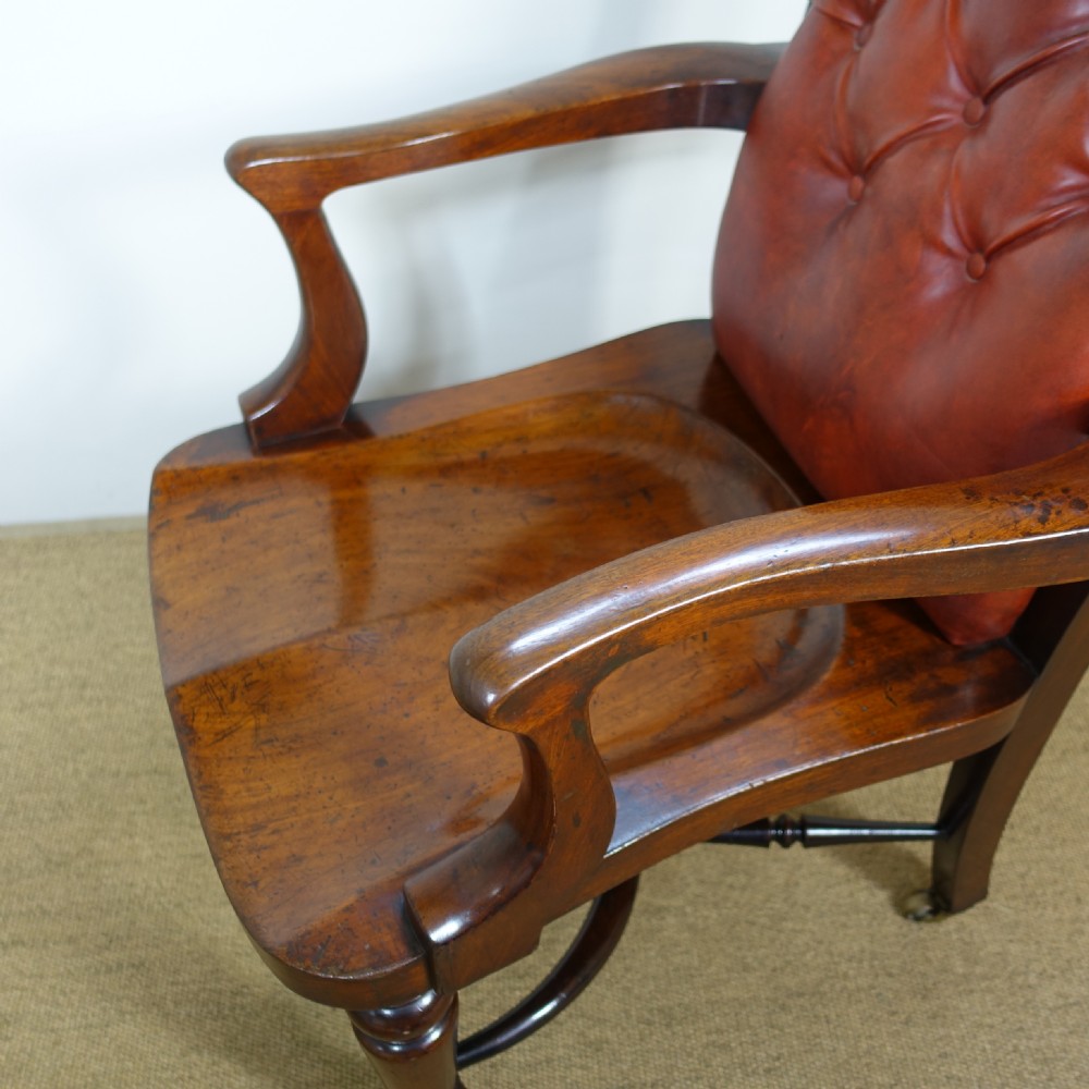 A 19th-century mahogany open armchair featuring a red buttoned-leather backrest, wooden seat, curved arms, turned front legs, and brass castors. View VI