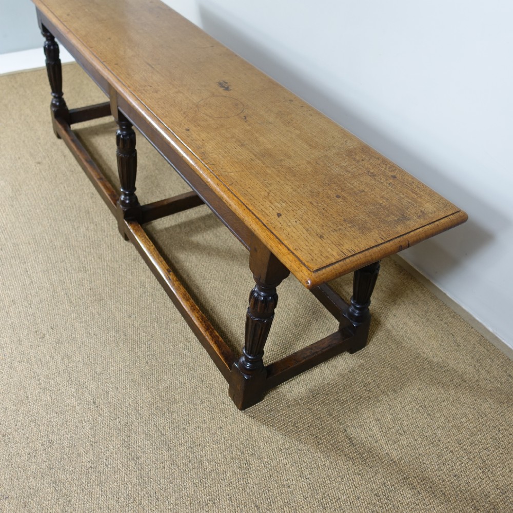 A long 19th-century dark oak stool featuring a flat plank top supported by six turned legs and low perimeter stretchers. View V